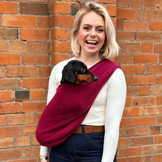 Woman holding a small dog in a red sling against a brick wall.