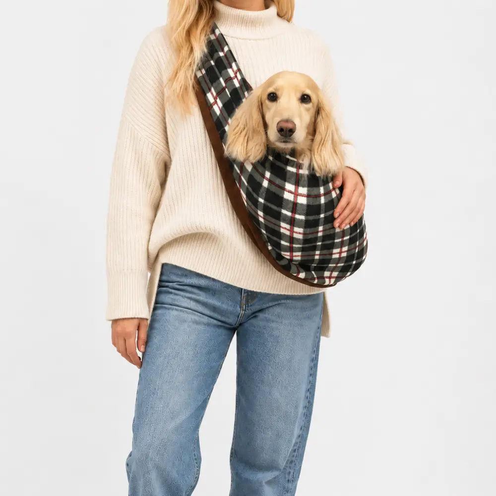 A woman wearing a cream knit sweater, blue jeans, and white sneakers carries a small golden dog in a black, white, and red plaid pet sling across her chest, set against a plain white studio background.